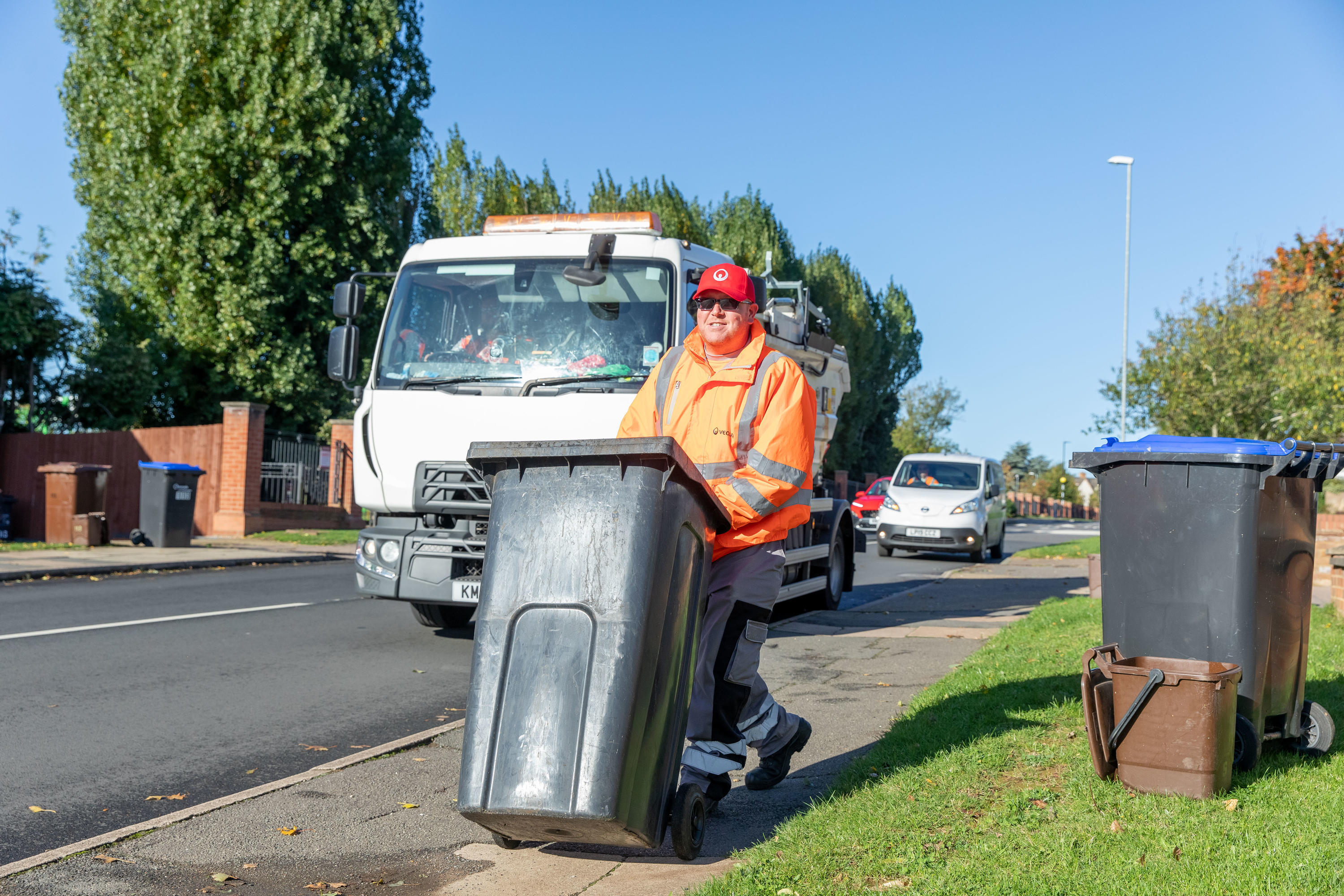 A loader wheels a bin down the street with a collection vehicle behind him.